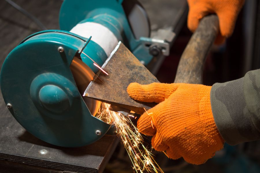 Hands in yellow gloves sharpening an axe blade using grinder